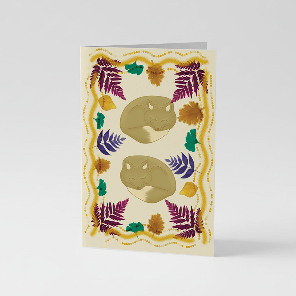 Greetings Card A5 - Illustration of two golden-brown foxes surrounded by burgundy and navy ferns, emerald leaves, golden flowers, and a delicate beaded border on cream background